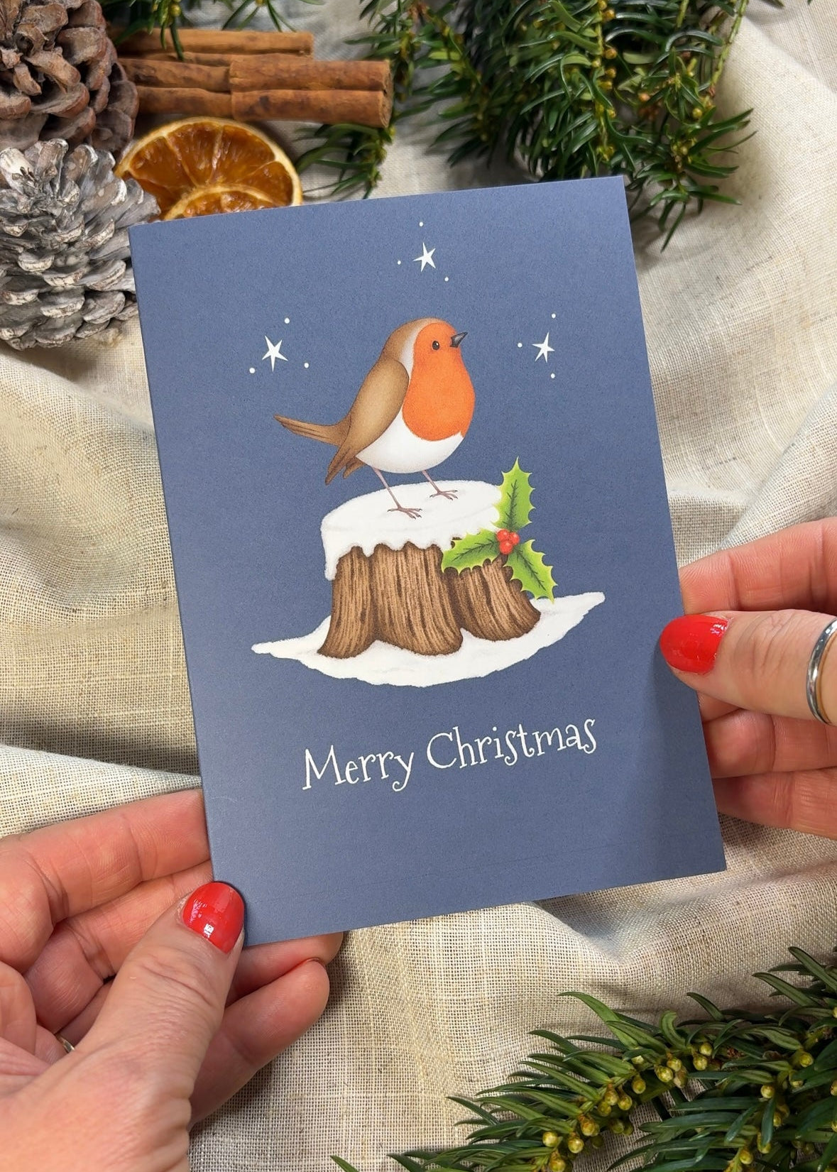 Christmas card with a robin on tree stump, held by hands with red nail polish, surrounded by festive decorations.
